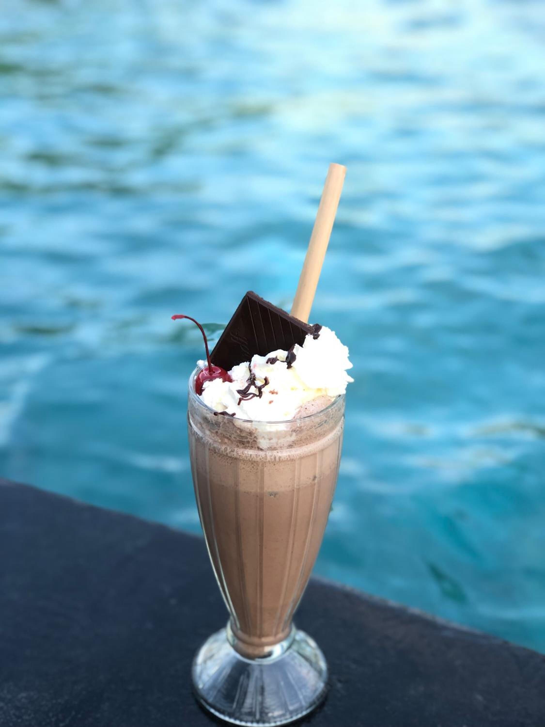 Un milkshake au chocolat, surmonté de crème fouettée et agrémenté d’un morceau de chocolat, est posé sur un verre, lui-même placé sur une table. En arrière-plan, on aperçoit un plan d’eau.