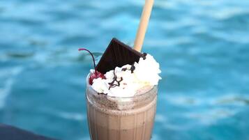 Un milkshake au chocolat, surmonté de crème fouettée et agrémenté d’un morceau de chocolat, est posé sur un verre, lui-même placé sur une table. En arrière-plan, on aperçoit un plan d’eau.