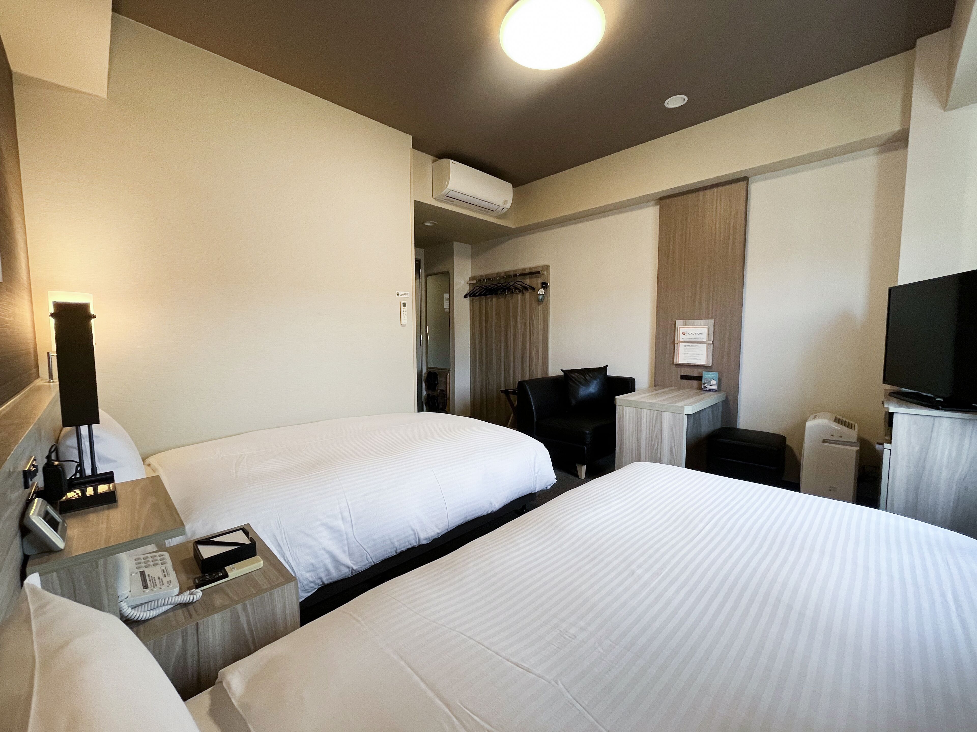 A hotel room with two beds, a desk with a telephone, a chair, a television, and an air conditioning unit.