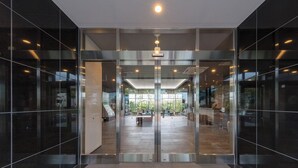Ingresso di un edificio moderno con porte in vetro, un lampadario centrale e vista su un'area interna con tavoli e sedie.