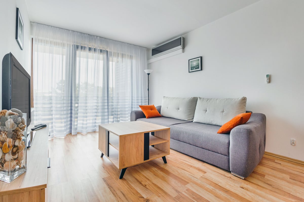 Ein modernes Wohnzimmer mit grauem Sofa, Couchtisch aus Holz und einem Fernseher links.