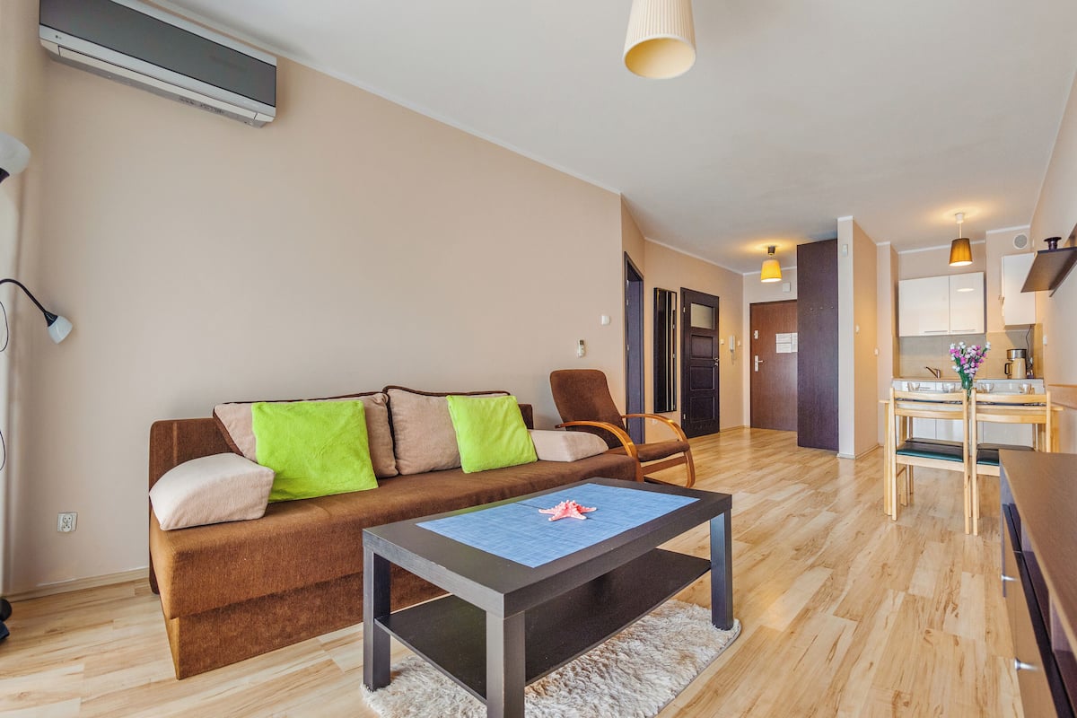Ein modernes Wohnzimmer mit einem braunen Sofa, einem Couchtisch und einem Fernseher. Im Hintergrund befindet sich ein Holzes Esstisch mit Stühlen und eine Küchenzeile.