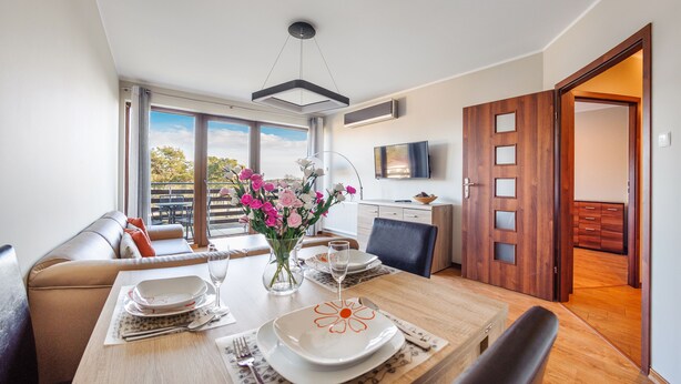 A dining area with a table set for two, a sofa, and a balcony with a view. - Apartamenty Sun & Snow Jasny Brzeg (Wladyslawowo)
