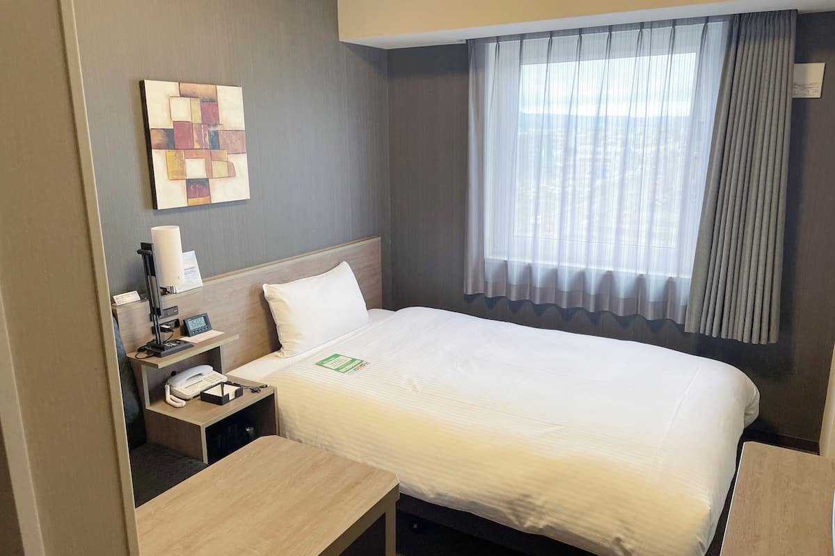 A hotel room with a bed, bedside table, lamp, and a window with curtains.