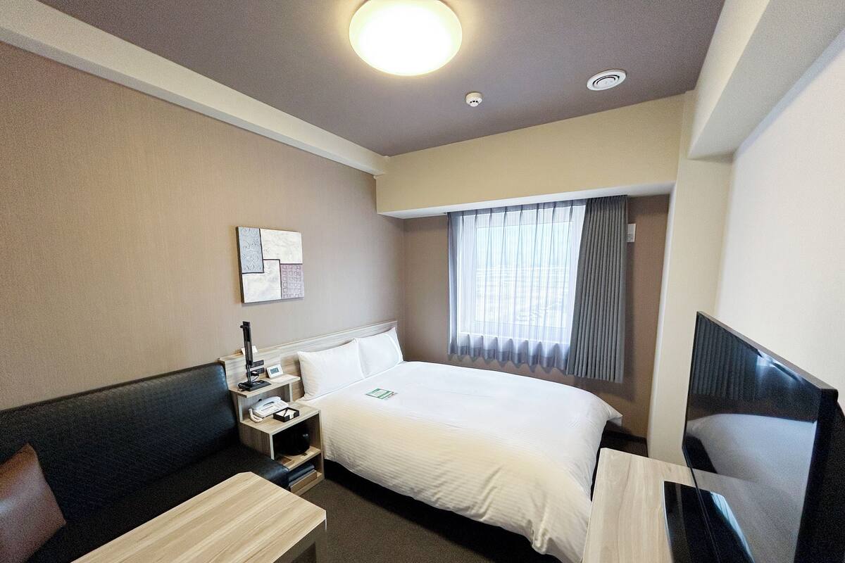 A hotel room with a bed, a desk, a chair, a TV, and a window with curtains.