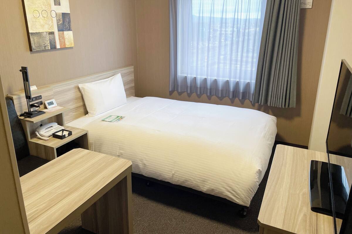A hotel room with a bed, a desk, a chair, a phone, a TV, and a window with curtains.