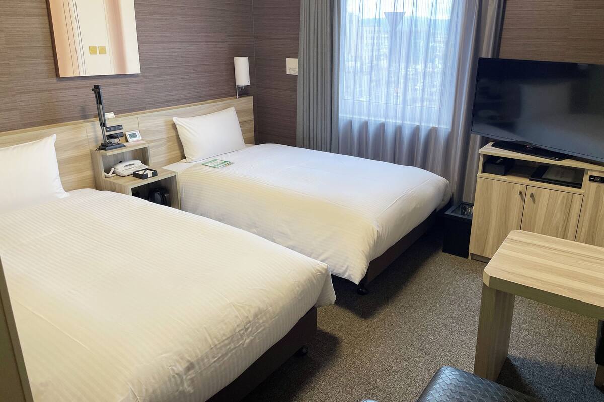 A hotel room with two beds, a TV, a desk, and a window with curtains.