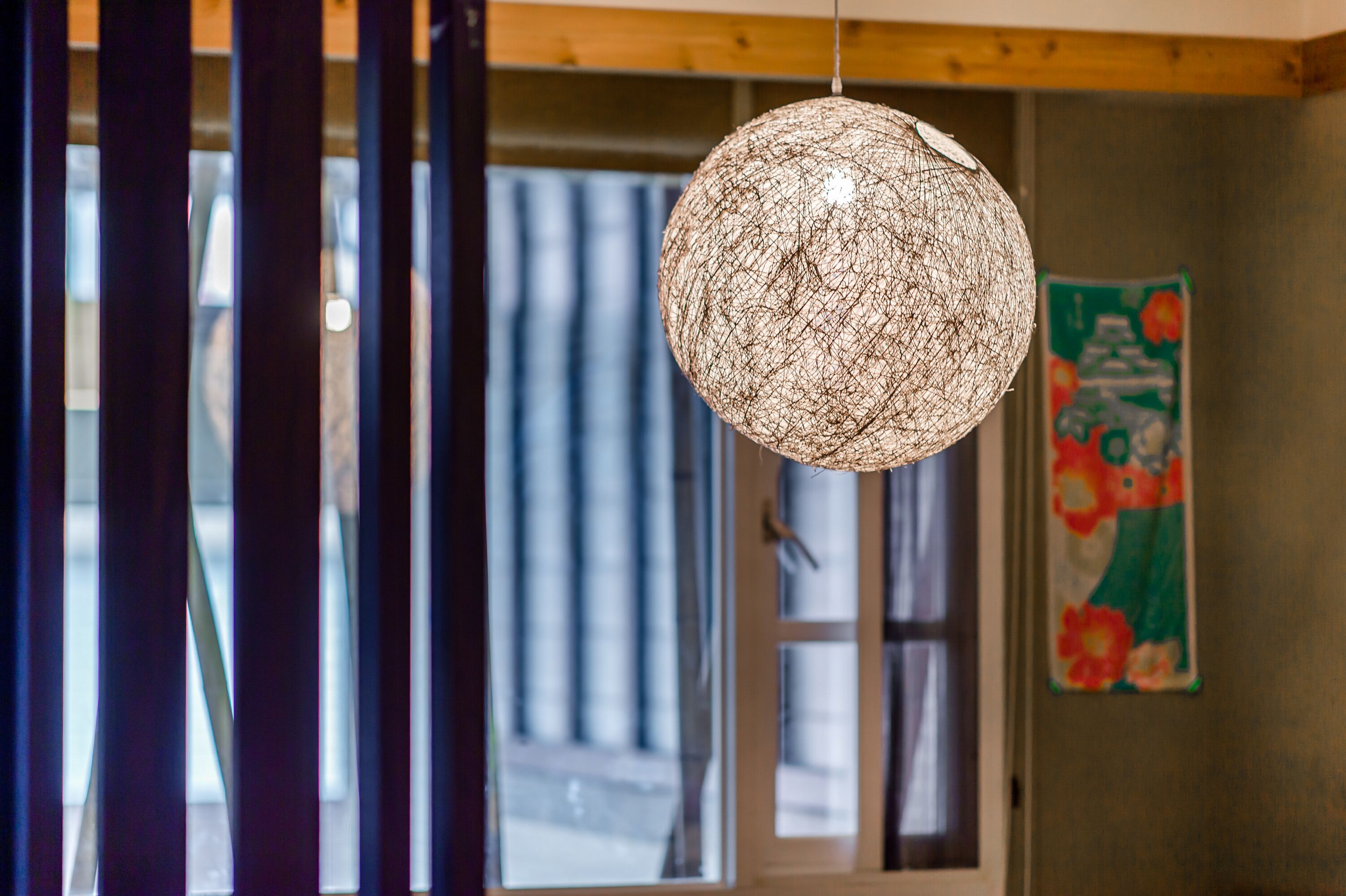 A spherical woven light fixture hanging from the ceiling in a room with wooden beams and a window with vertical blinds.