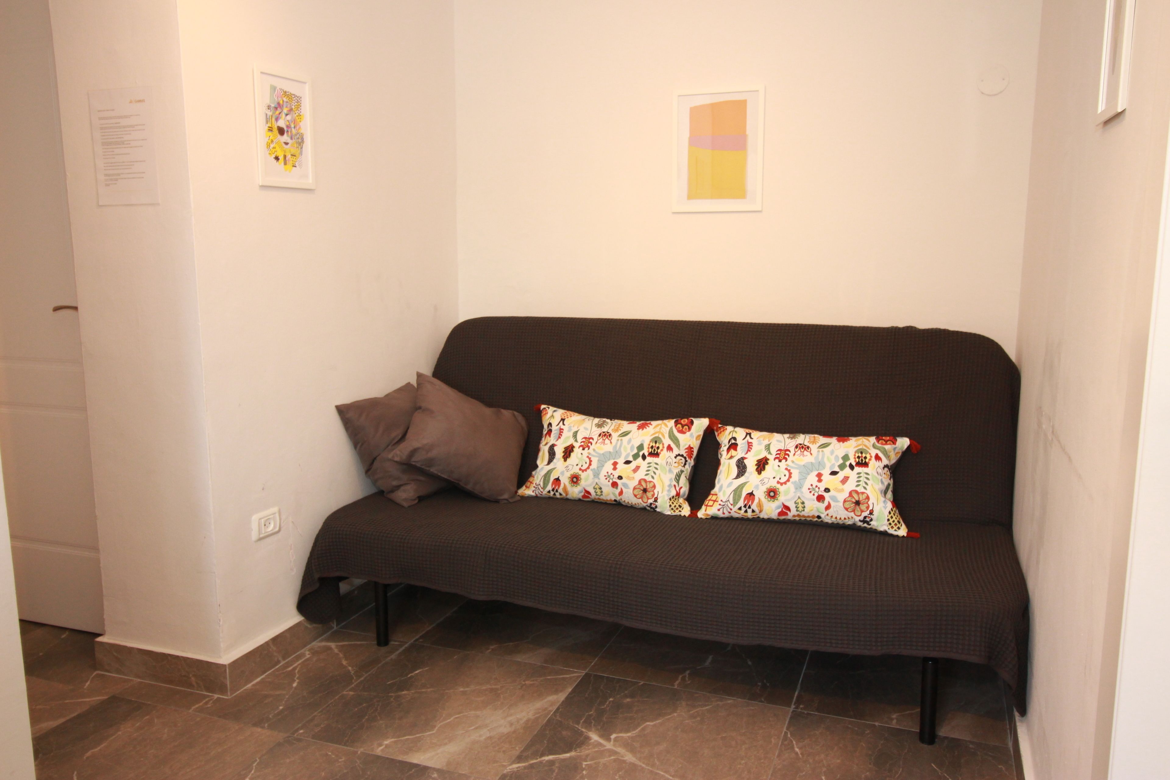 Un sofá marrón oscuro con cojines estampados, una pared blanca con dos cuadros enmarcados y un suelo de baldosas.