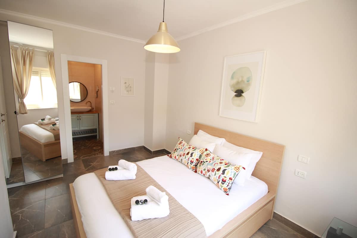 A hotel room with a bed, a bedside table, a mirror, a window with curtains, and a wall-mounted light fixture.