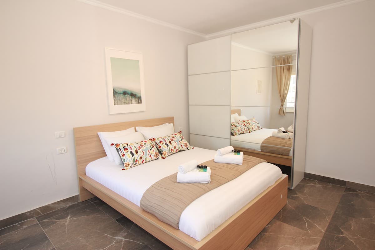 A neatly made bed with a wooden headboard, a large mirror, and a framed picture on the wall.