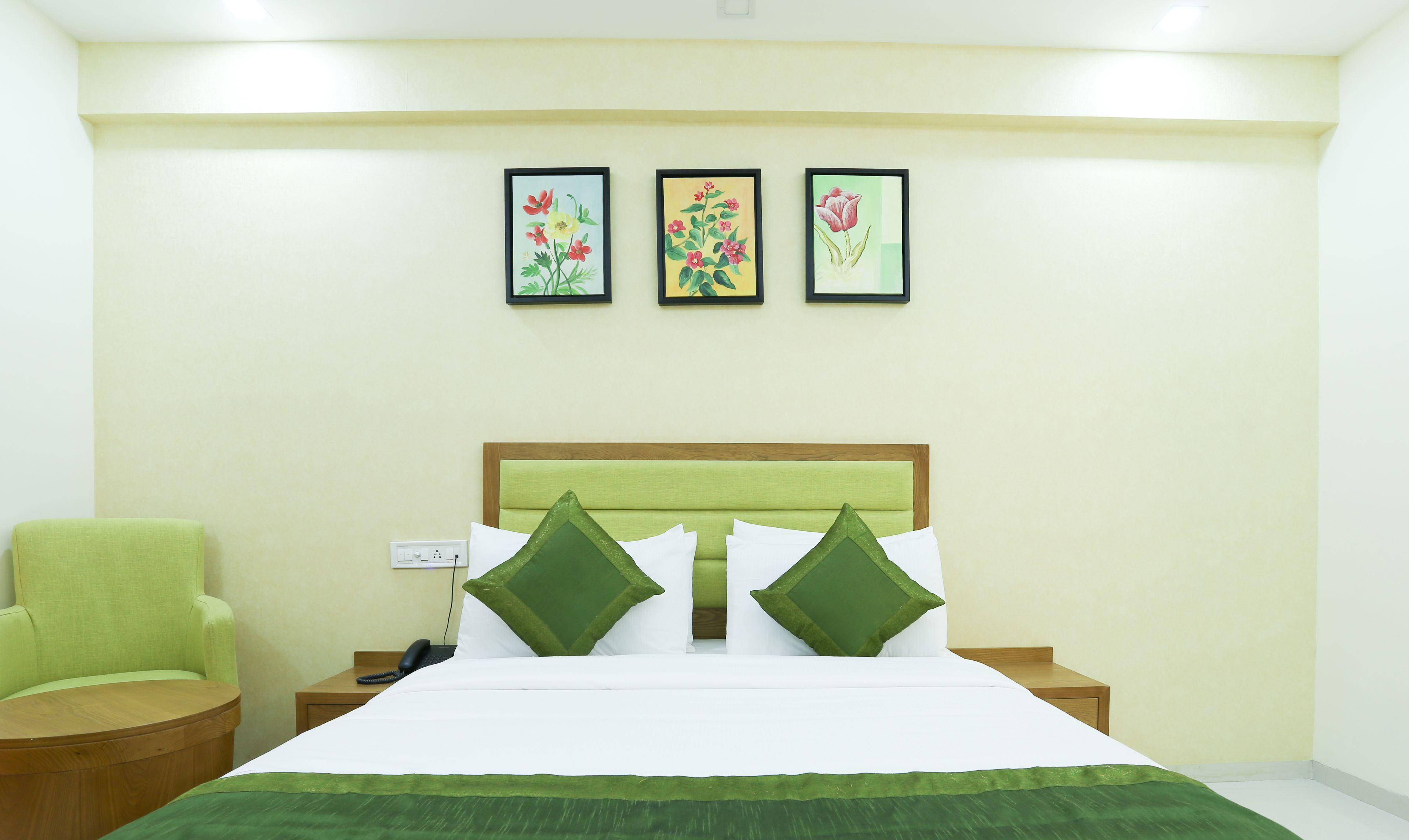 A hotel room with a bed, green pillows, a green chair, and three framed floral paintings on the wall.