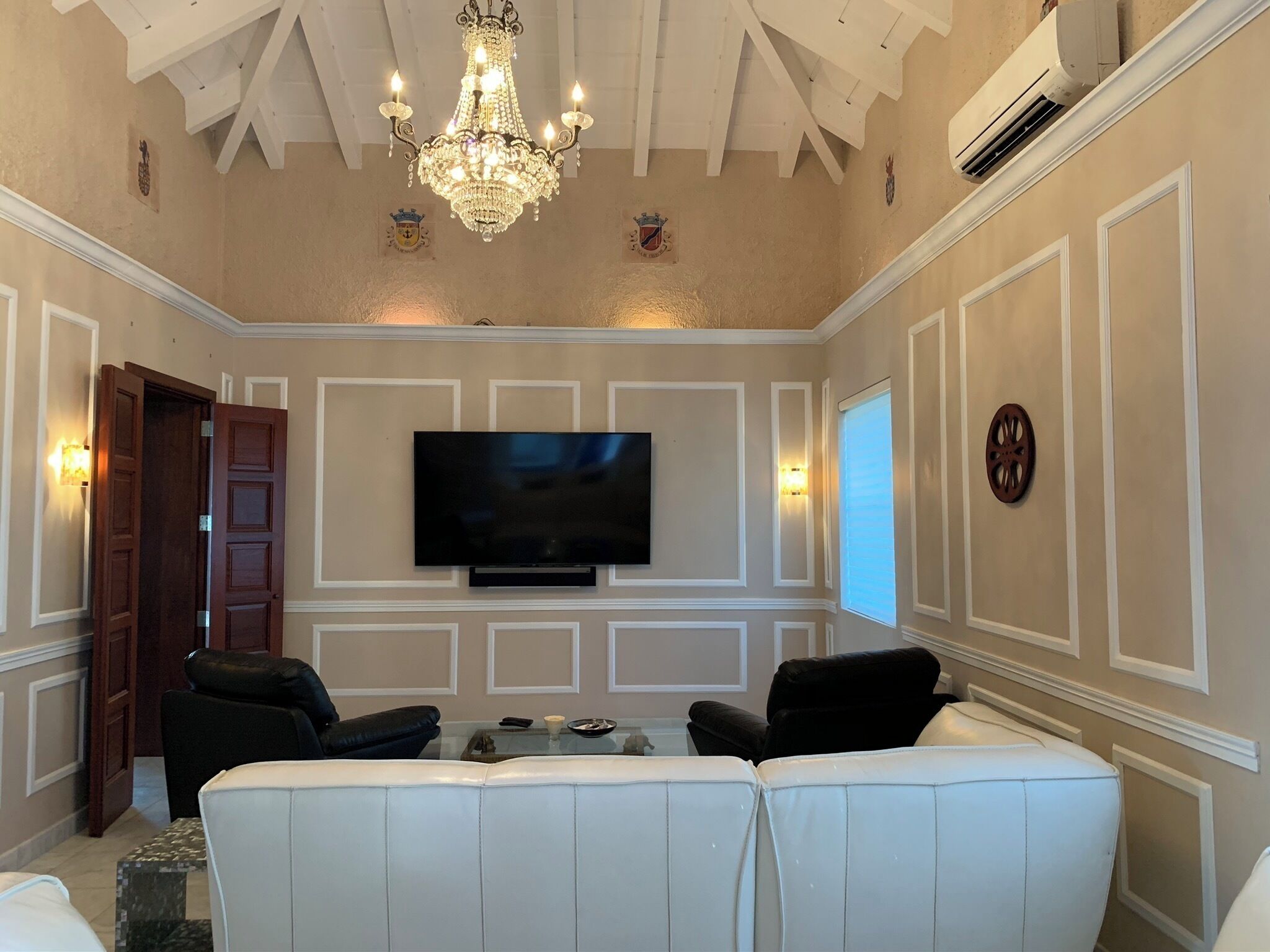 A living room with a large flat-screen TV, a white leather sofa, a chandelier, and a wooden door.