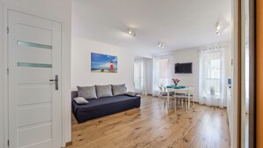 A room with a wooden floor, a dark sofa with cushions, a dining table with chairs, a TV mounted on the wall, and a painting of a lighthouse.