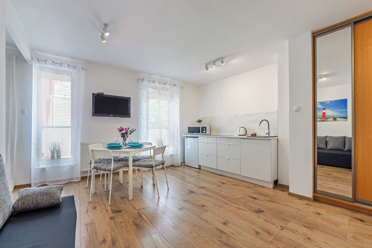 A modern kitchen with white cabinets, a microwave, and a sink. There is a dining table with chairs and a TV mounted on the wall.
