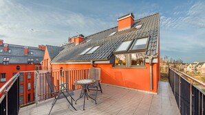 A rooftop terrace with a table and chairs, a view of the city, and a red building with a grey roof.