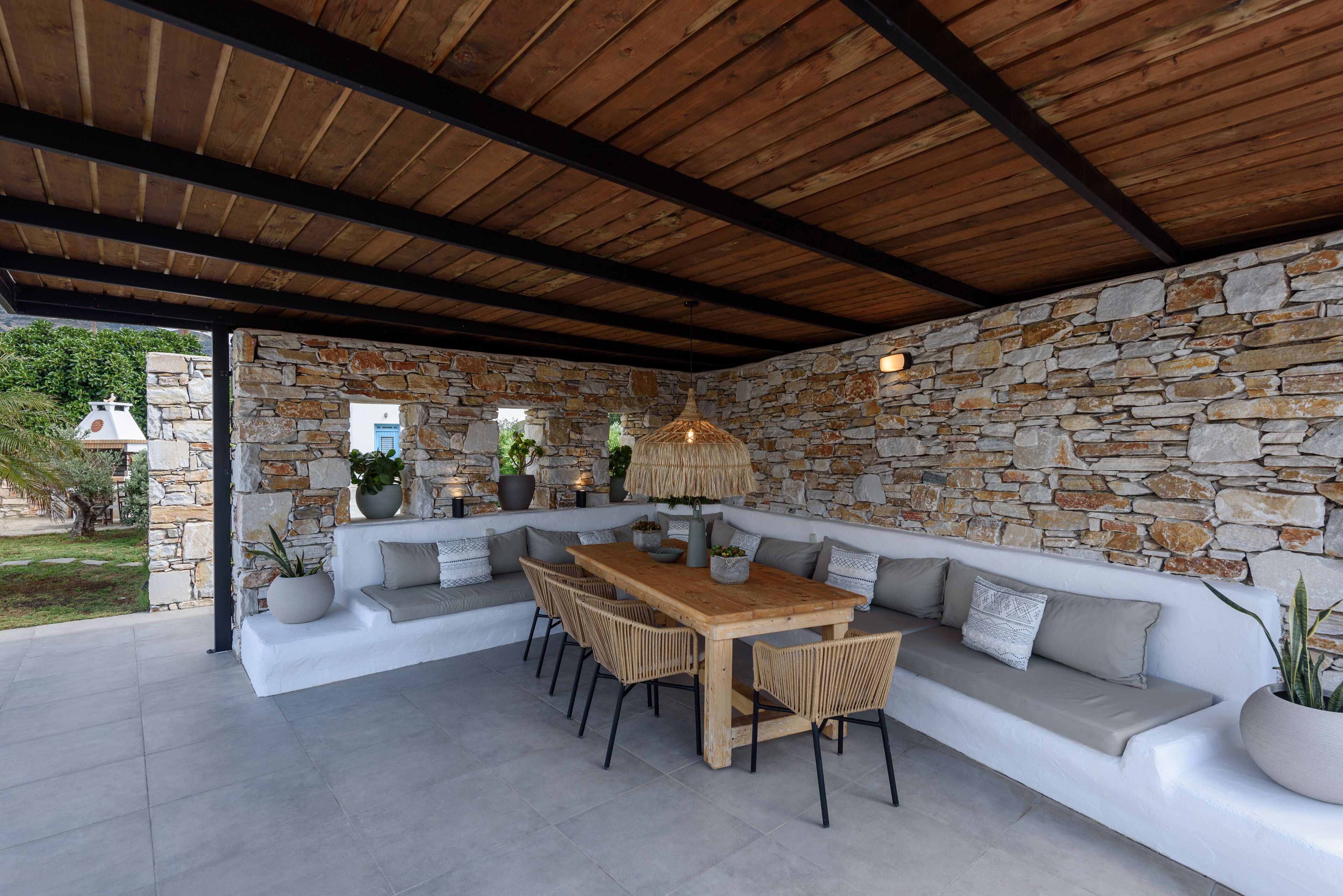 Un área de estar al aire libre con techo, paredes de piedra, una mesa de madera y sillas.