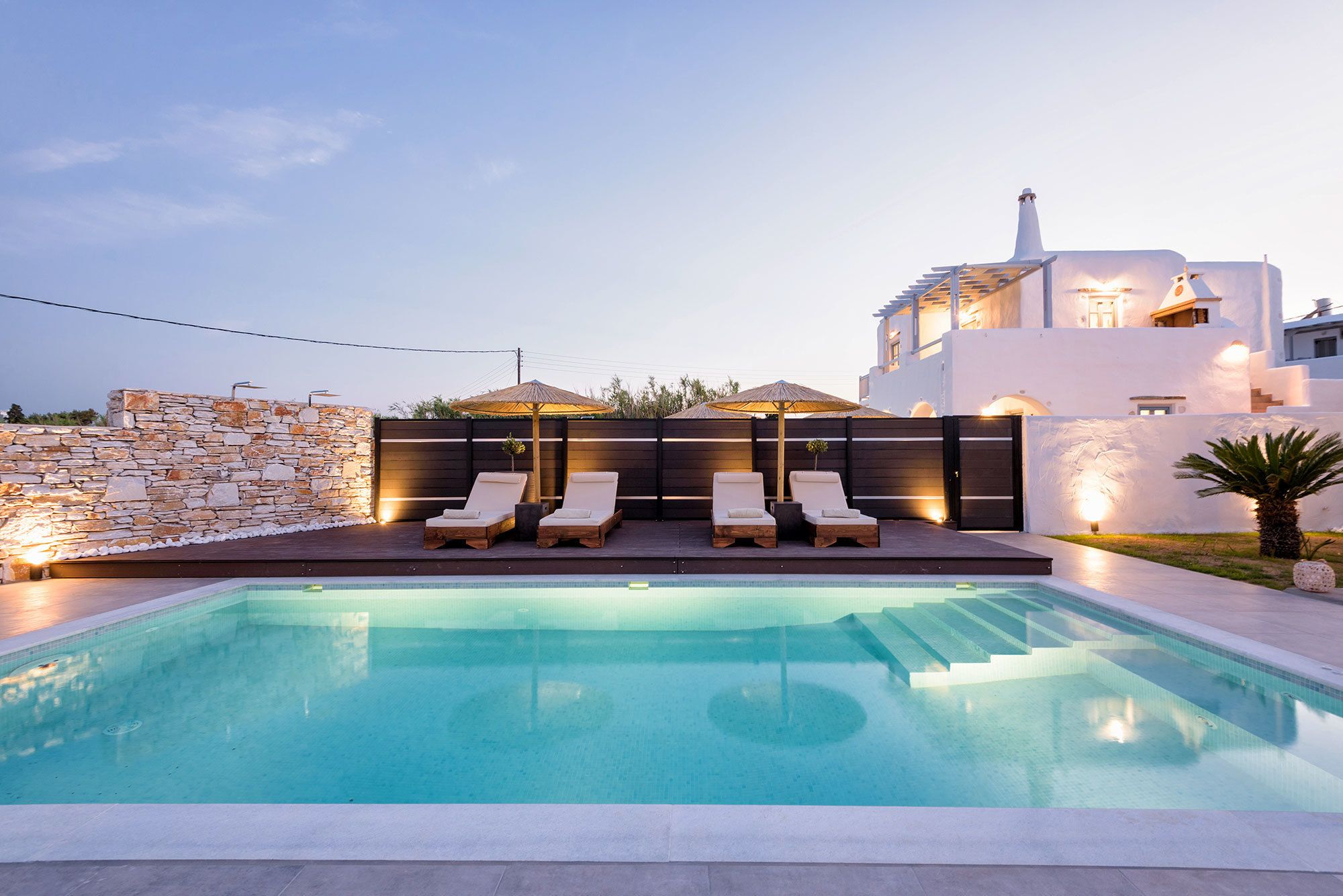 Piscina com espreguiçadeiras e guarda-sóis, cercada por um muro de pedra e um edifício branco com chaminé.