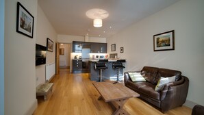 A modern living room with a wooden floor, a brown leather sofa, a wooden coffee table, and a kitchen in the background. - Cs Serviced Apartments (Ulverston)