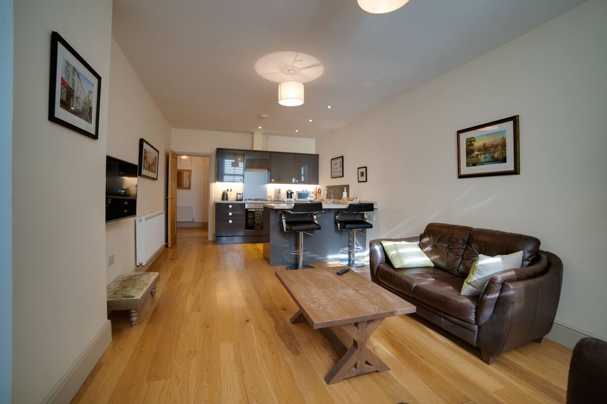 A modern living room with a wooden floor, a brown leather sofa, a wooden coffee table, and a kitchen in the background.