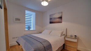 A bedroom with a bed, bedside table, and a window with blinds. - Cs Serviced Apartments (Ulverston)