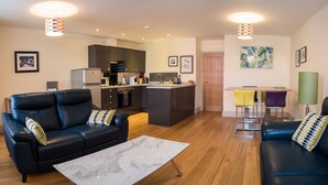 A modern living room with a sofa, coffee table, and a kitchen area with a dining table. - Cs Serviced Apartments (Ulverston)