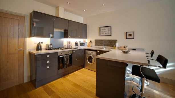 A modern kitchen with grey cabinets, a built-in oven, microwave, and washing machine. There is a bar area with two black bar stools and a small island with a sink. - Cs Serviced Apartments (Ulverston)