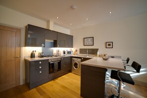 A modern kitchen with grey cabinets, a built-in oven, microwave, and washing machine. There is a bar area with two black bar stools and a small island with a sink.