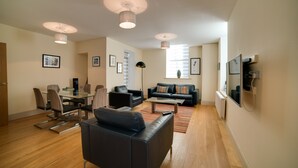A modern living room with a dining area, a sofa, a coffee table, and a TV mounted on the wall. - Cs Serviced Apartments (Ulverston)