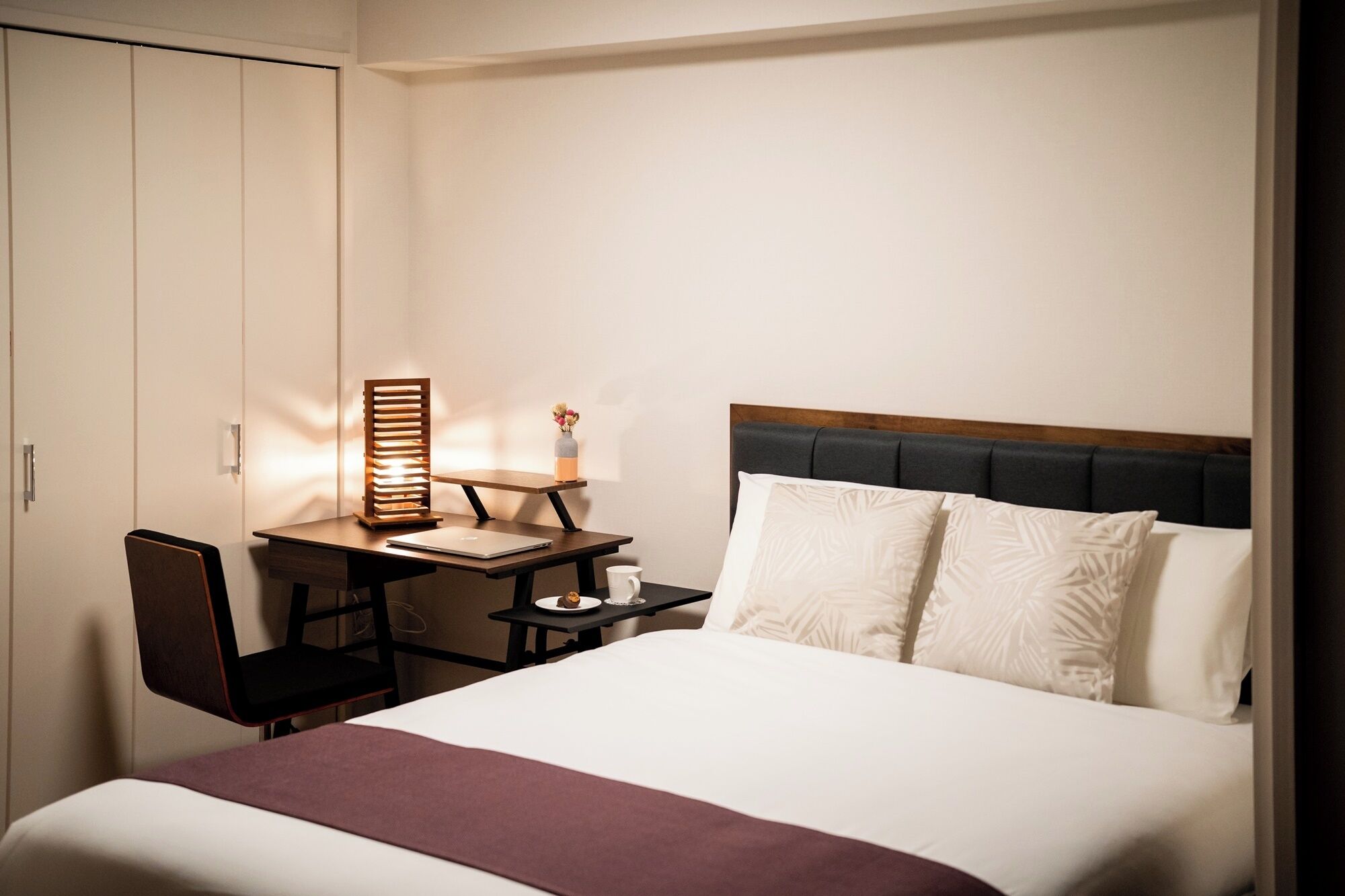 A hotel room with a bed, a desk with a chair, a nightstand, and a wardrobe.