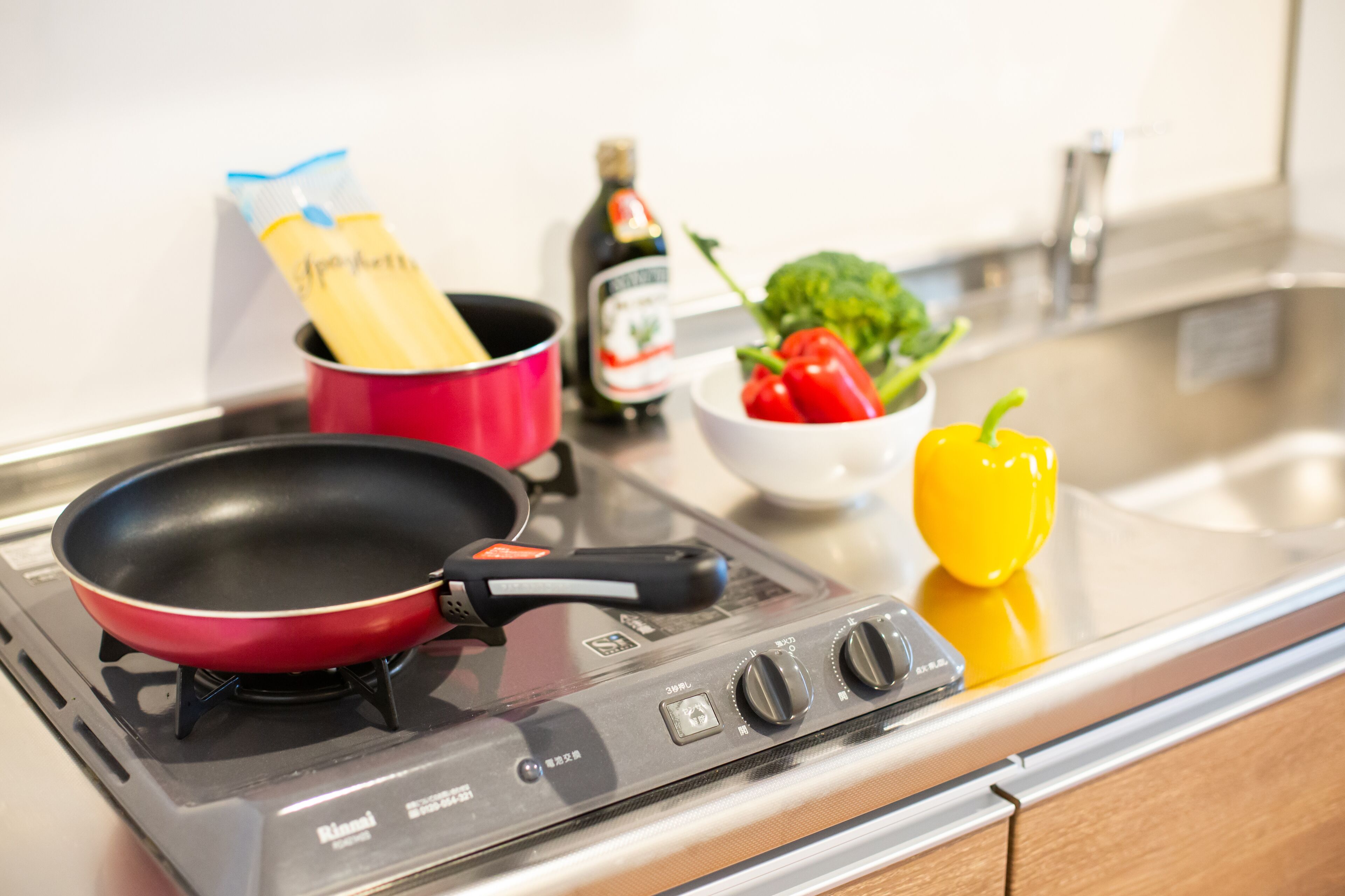 A kitchen with a red frying pan on a stove, a pot, a bottle of olive oil, a bowl of red peppers, and a yellow bell pepper.