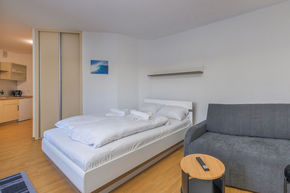A modern hotel room with a bed, a grey armchair, a small wooden table, and a painting on the wall.