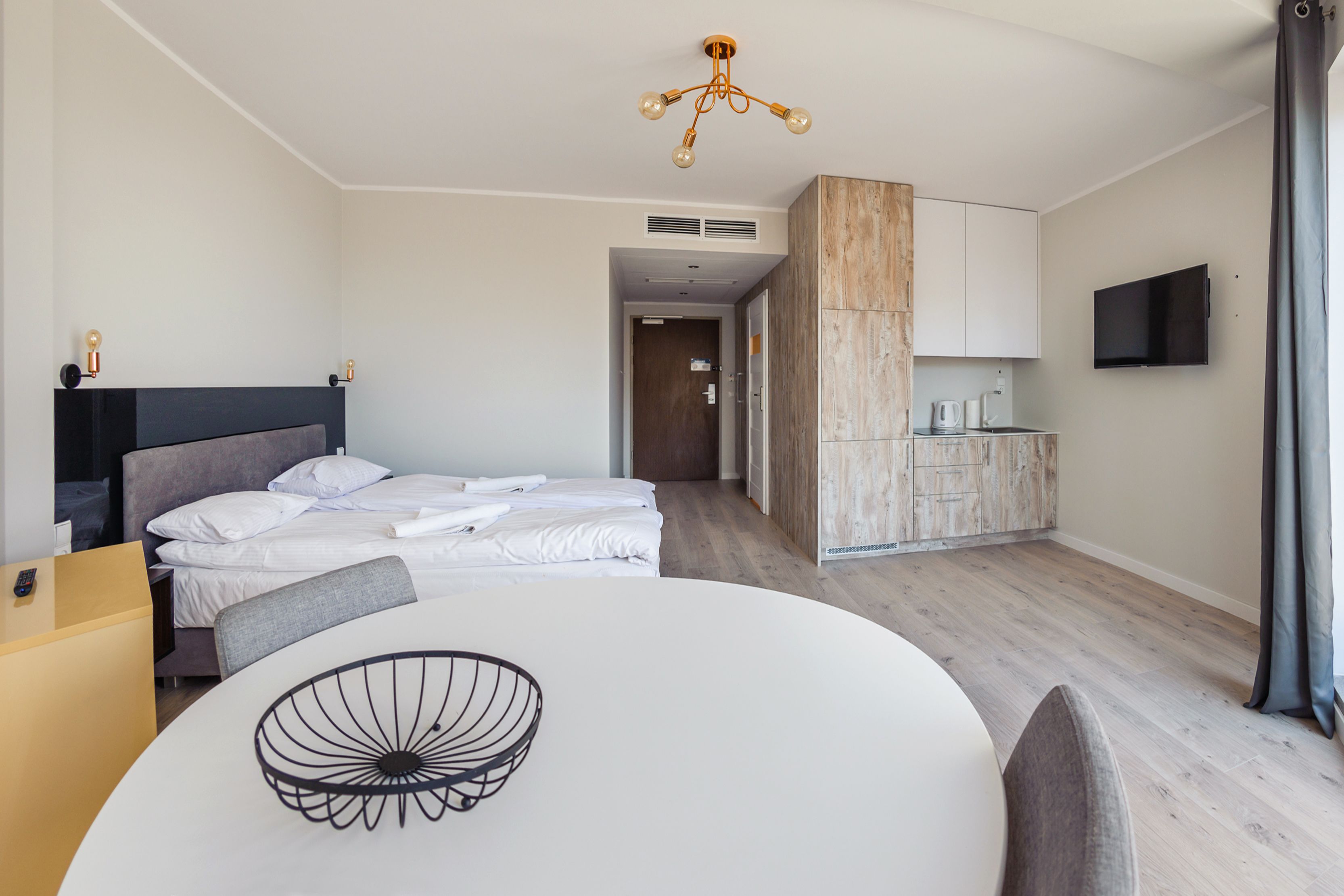 A modern hotel room with a bed, a dining table, a kitchen area, and a TV.