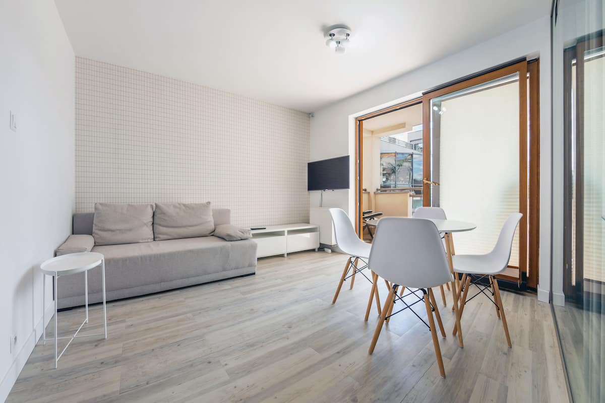 A modern living room with a grey sofa, a small white table, and a dining area with white chairs and a glass table.