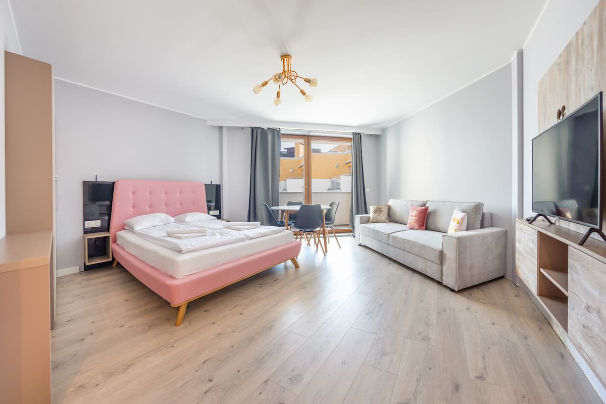 A modern hotel room with a pink bed, a grey sofa, a TV, and a balcony with curtains.