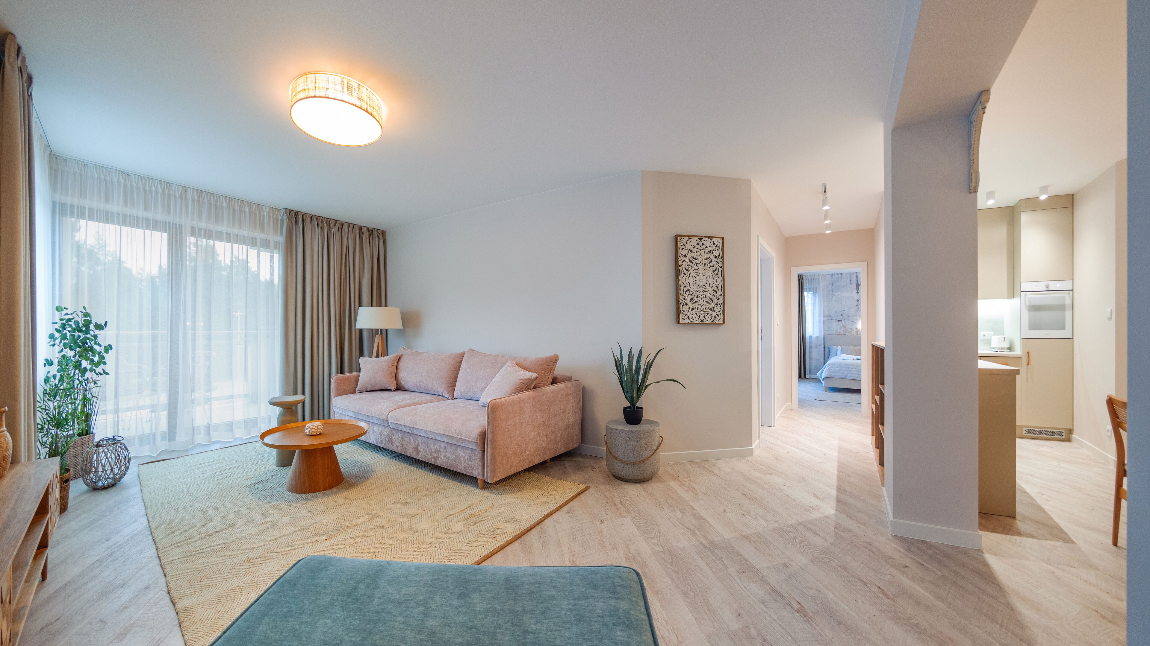 En moderne stue med sofa, sofabord og en potteplante. Der er kig til et køkkenområde gennem en åben dør.