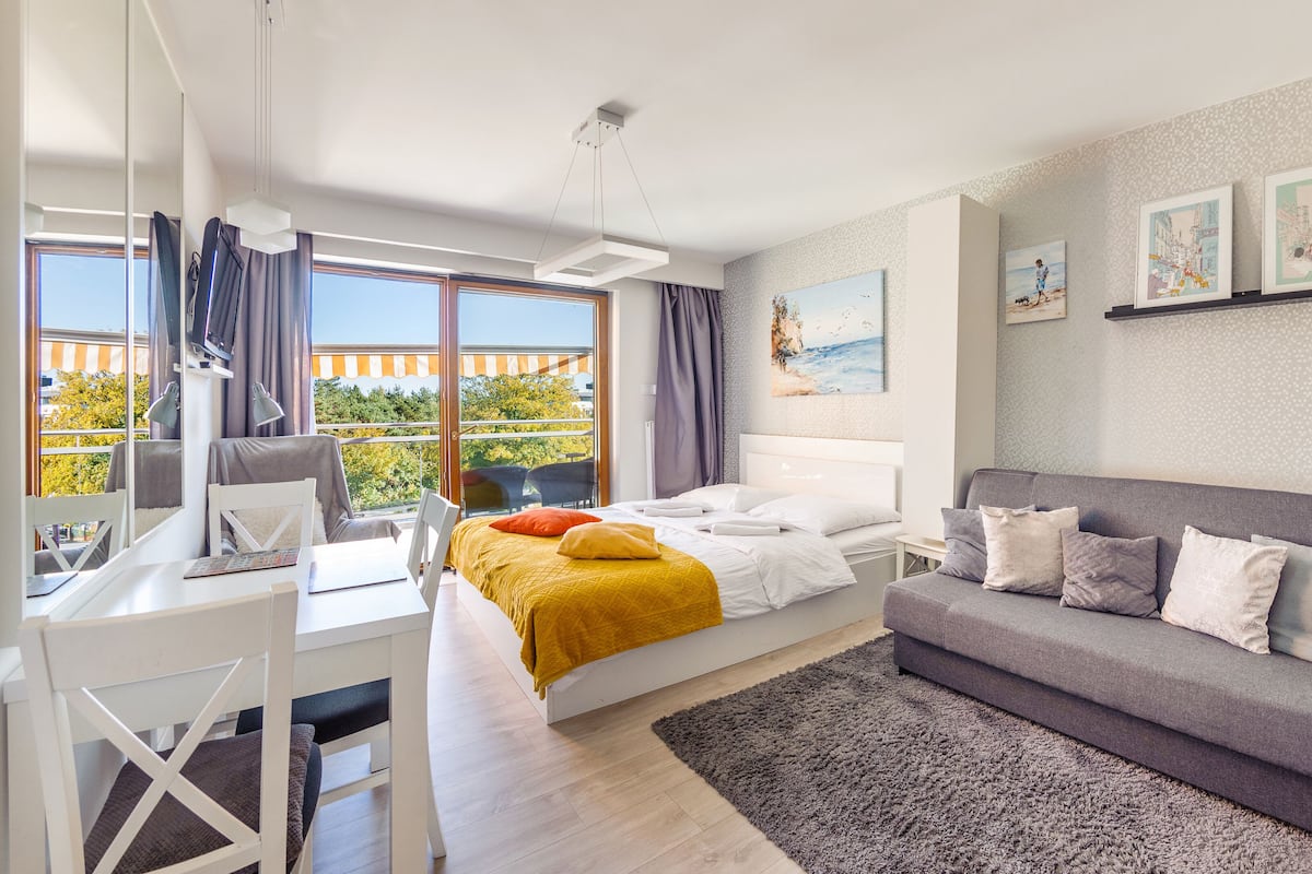 A modern bedroom with a bed, a grey sofa, a desk with a chair, and a balcony with curtains.