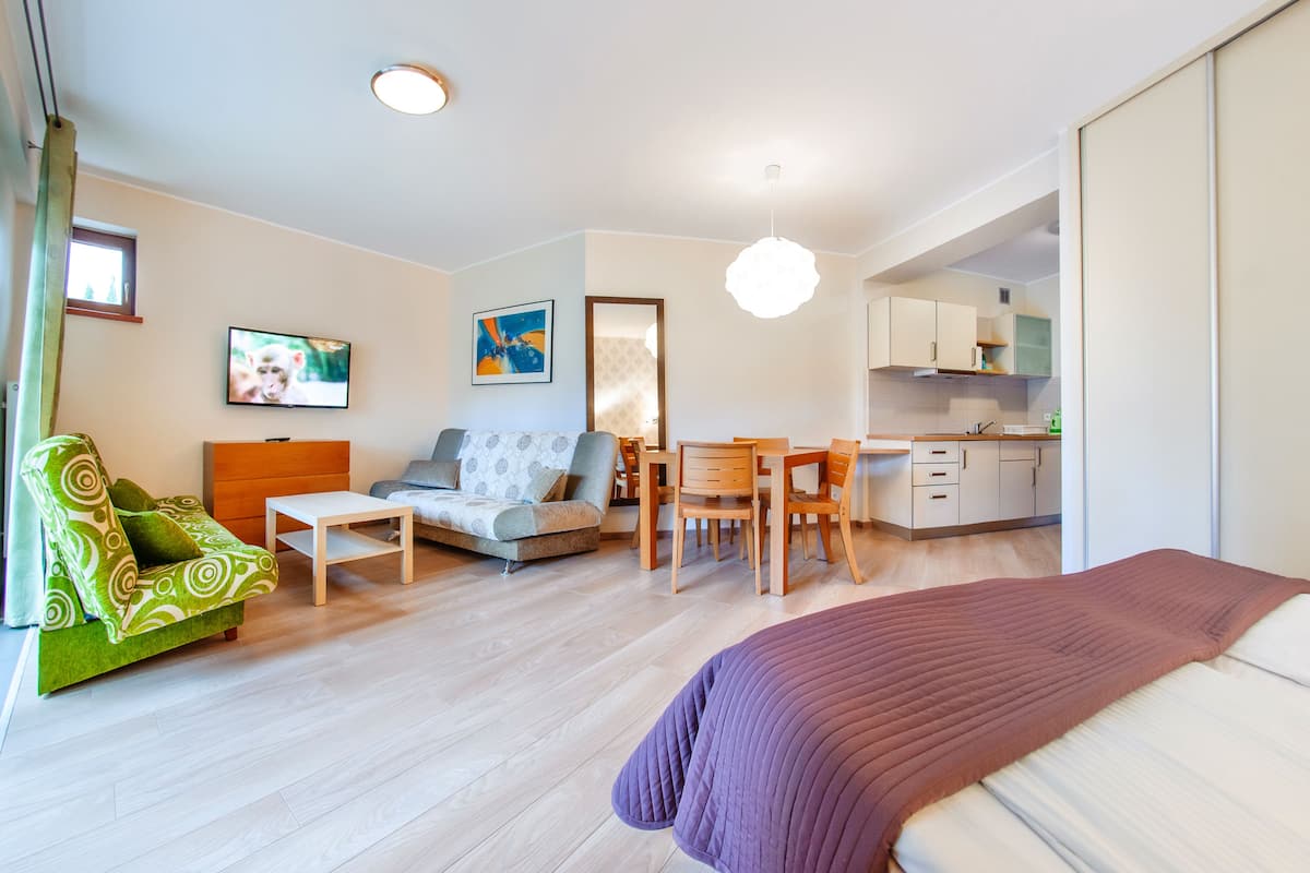 A modern hotel room with a bed, a sofa, a dining table, and a kitchen area.