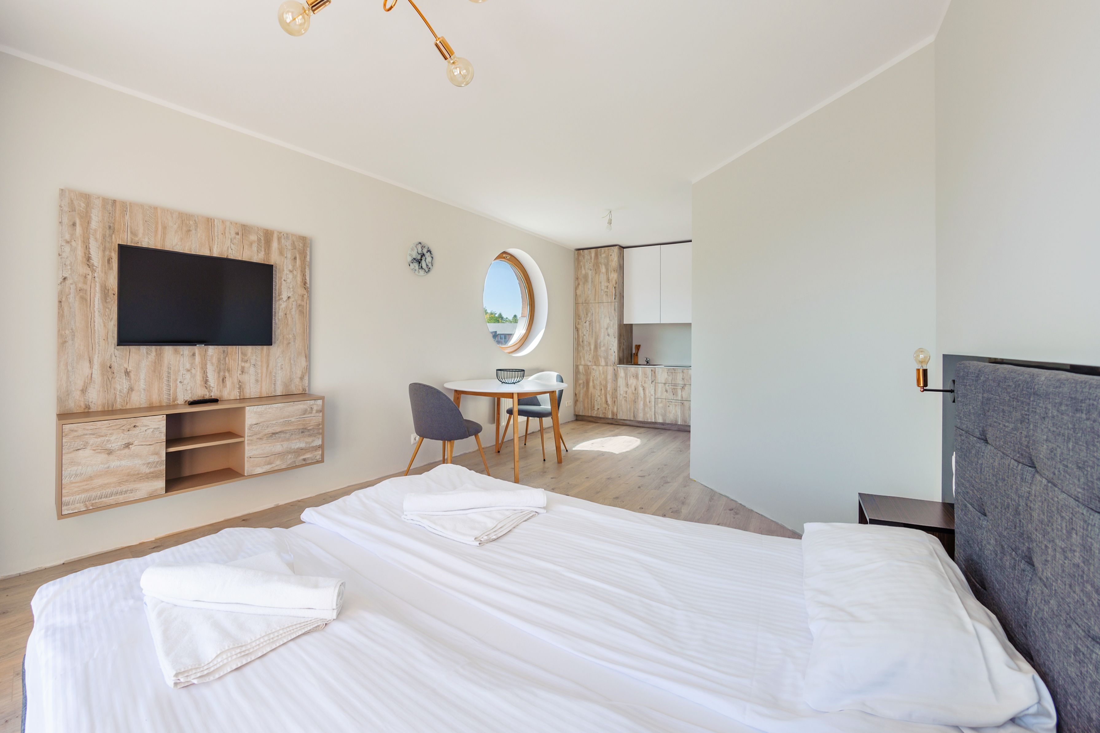 Une chambre d’hôtel moderne avec un grand lit, un bureau, une chaise, un téléviseur fixé sur un panneau en bois et un miroir rond.
