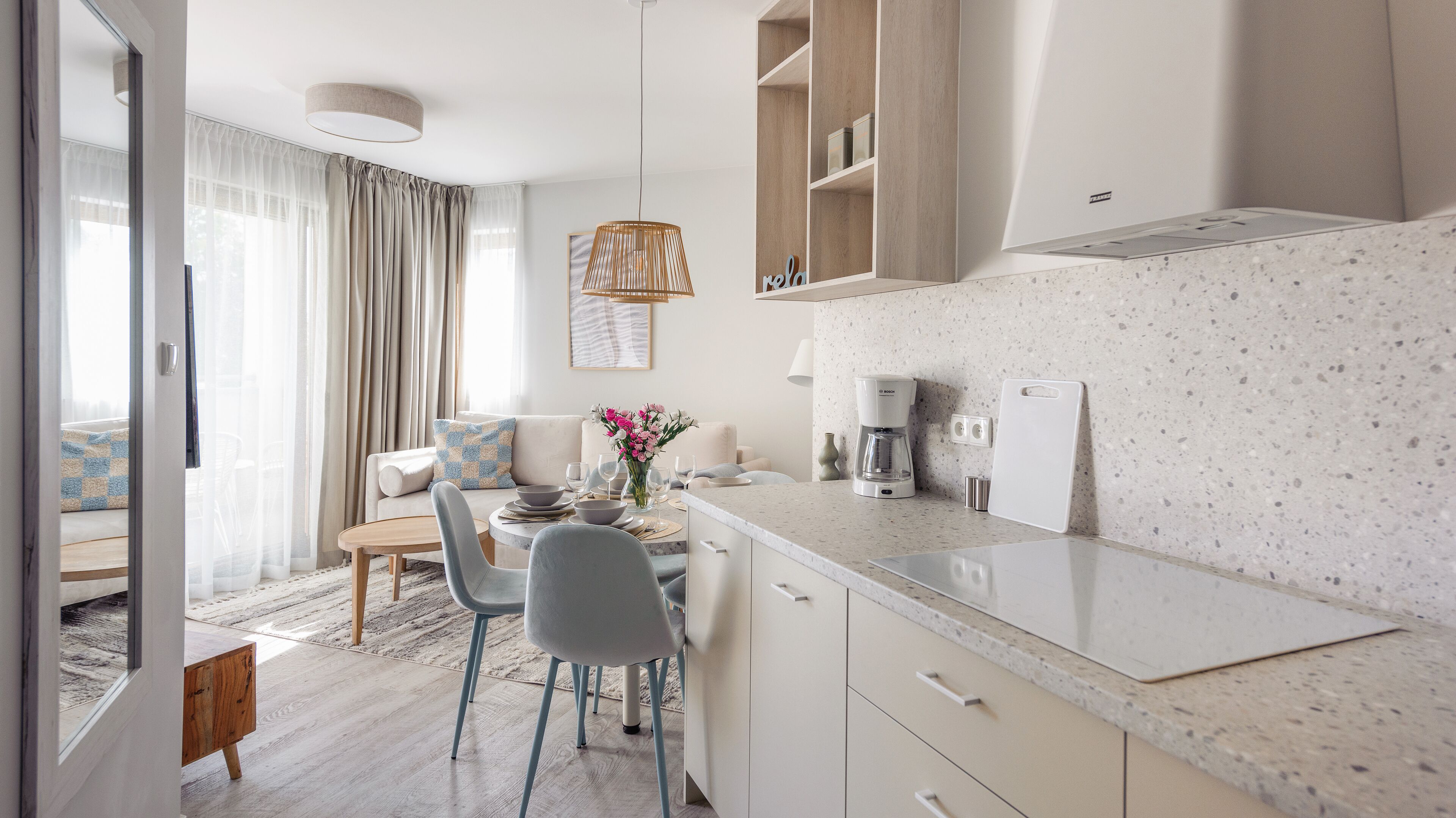 A modern kitchen with a coffee maker, a dining area with a table and chairs, and a living area with a sofa and curtains.