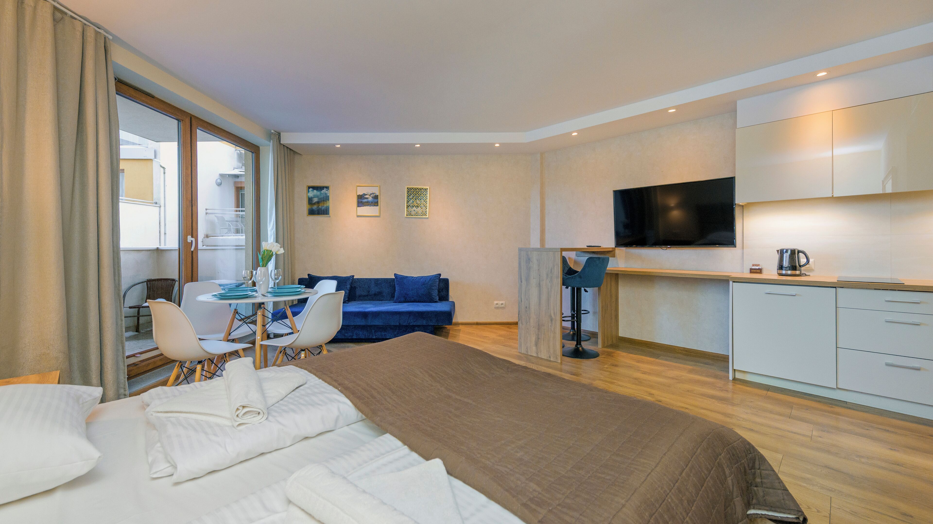 A modern hotel room with a large bed, a dining area with a table and chairs, a blue sofa, a kitchen area with a TV, and a view of the outside balcony.