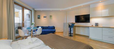 A modern hotel room with a large bed, a dining area with a table and chairs, a blue sofa, a kitchen area with a TV, and a view of the outside balcony.