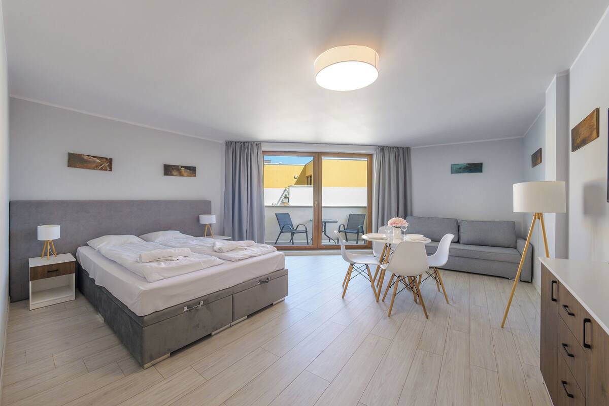 A modern hotel room with a bed, a dining area with a table and chairs, a sofa, and a TV.