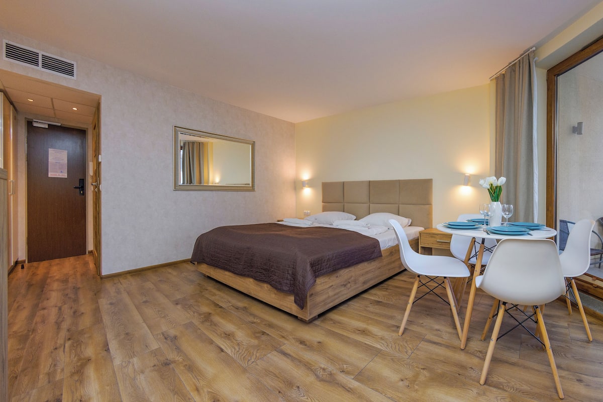 A hotel room with a large bed, a dining table with chairs, a mirror, and a window with curtains.