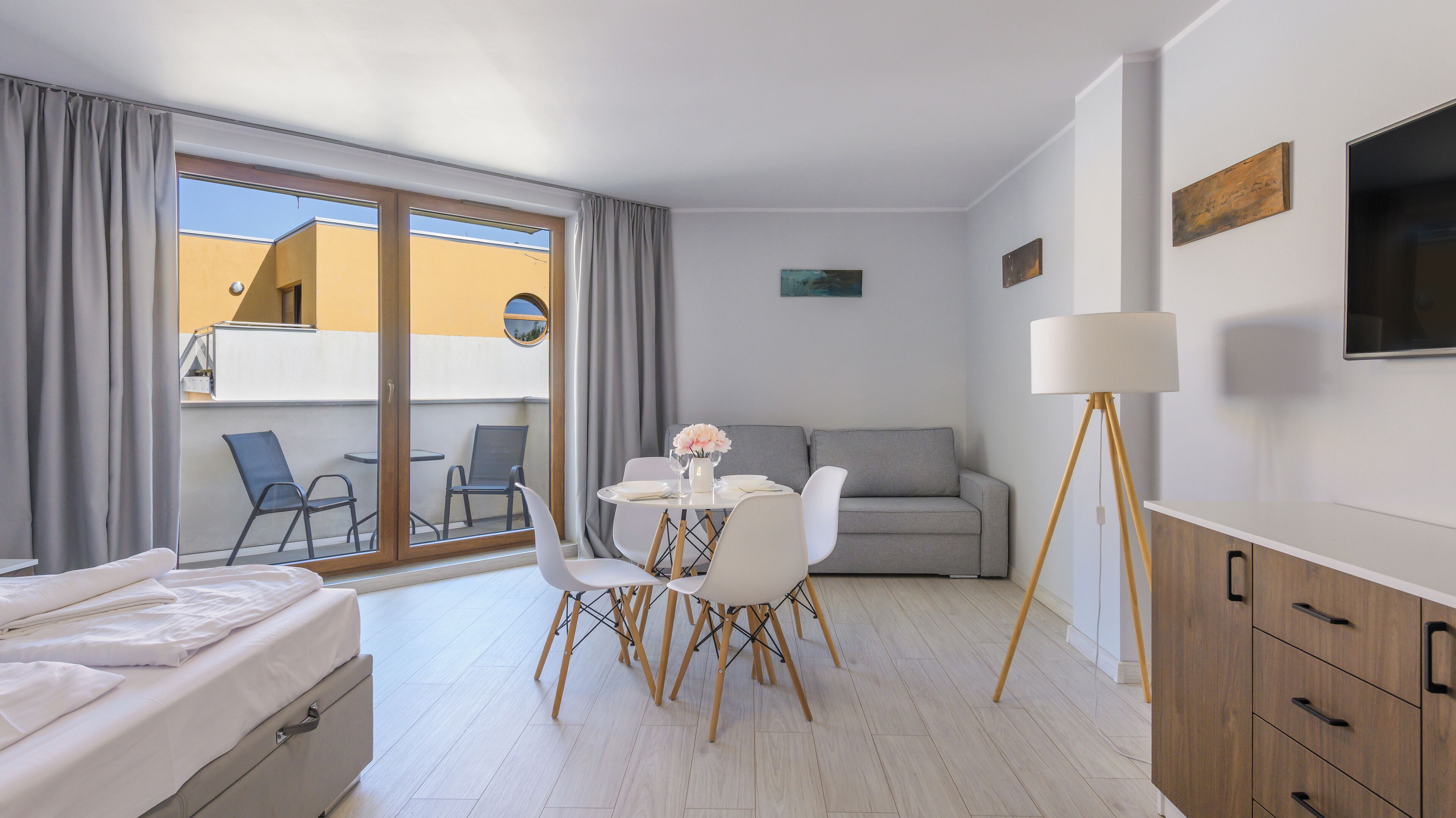 A modern hotel room with a balcony, a dining table with chairs, a sofa, a floor lamp, and wooden cabinets.