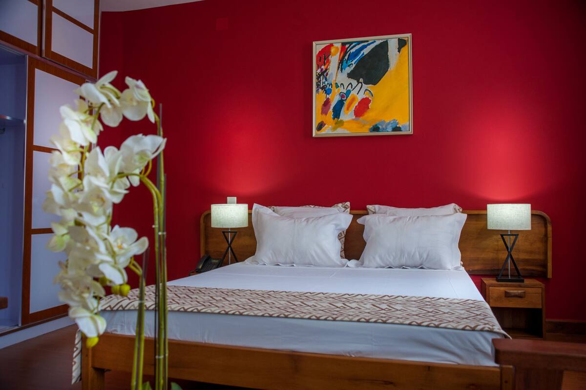 A hotel room with a large bed, two bedside tables, a red wall, and a painting above the bed.