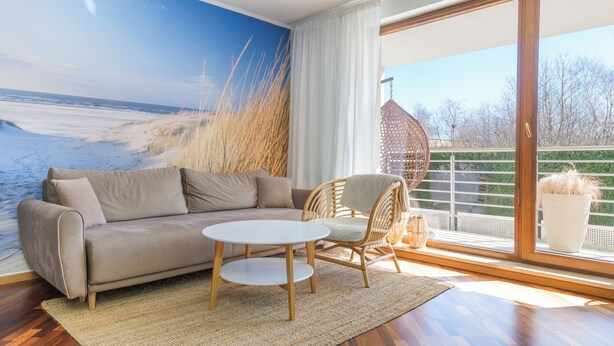 A living room with a sofa, a small table, a wicker chair, a large window with a view of the beach, and a mural of the beach. - Apartamenty Sun&Snow Na Klifie (Wladyslawowo)