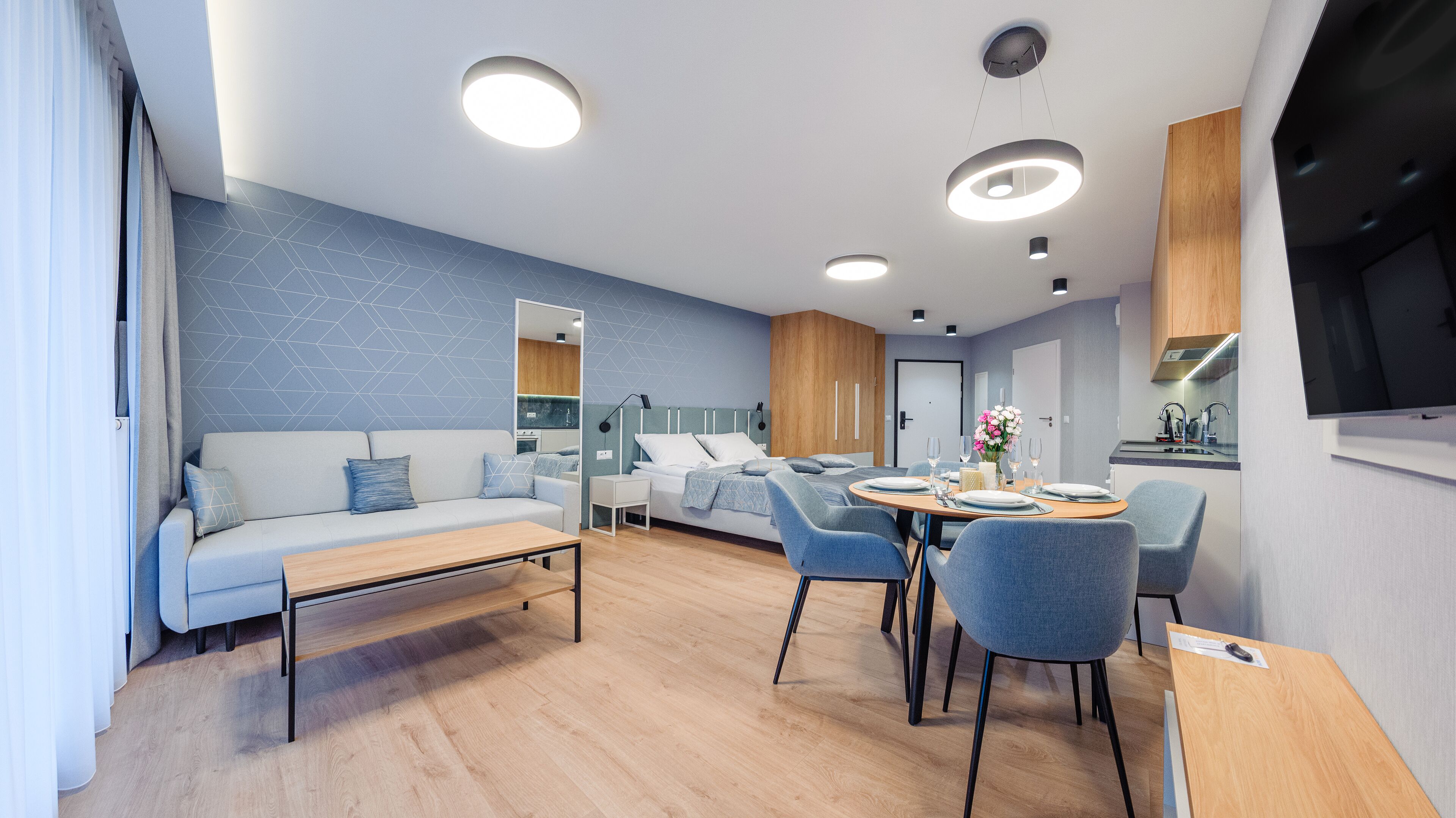 Uma sala de estar moderna e bem iluminada, com sofá, mesa de jantar e cadeiras. Há uma área de cozinha com armários e uma TV fixada na parede.