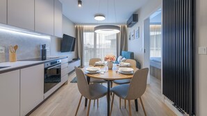 A modern dining area with a round table set for four, a kitchen with a built-in oven, and a large window with curtains. - Apartamenty Sun&Snow Na Klifie (Wladyslawowo)