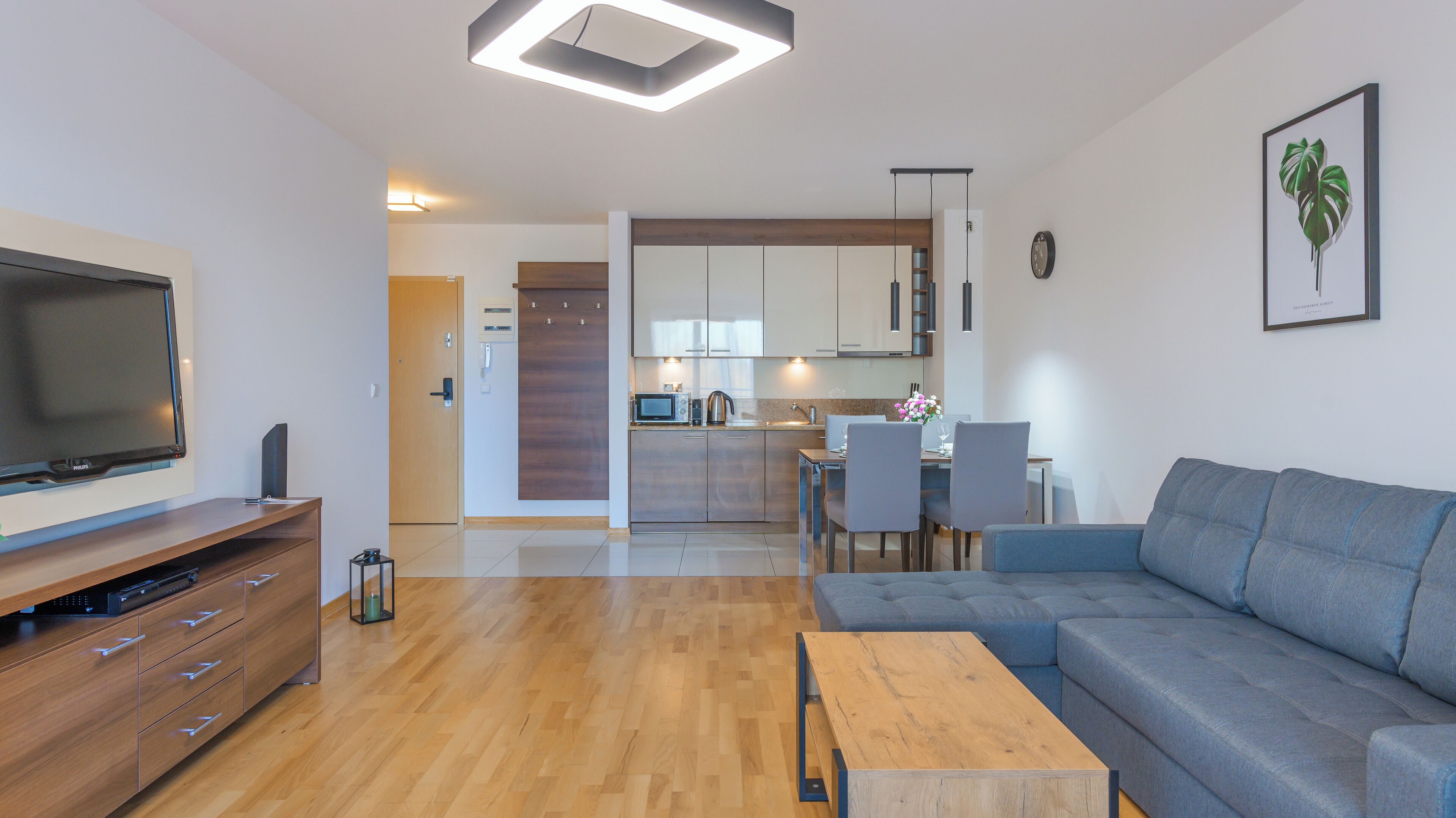 A modern living room with a flat-screen TV, wooden furniture, a grey sofa, and a dining area with a table and chairs.